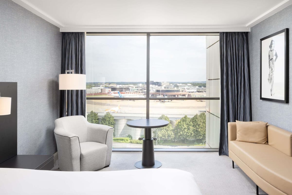 A hotel room with a large window, a bed, a chair, a table, and a sofa.