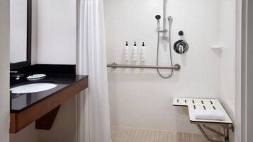Baño con lavamanos sobre encimera negra, azulejos blancos, ducha con cabezal de ducha manual y barra de apoyo.