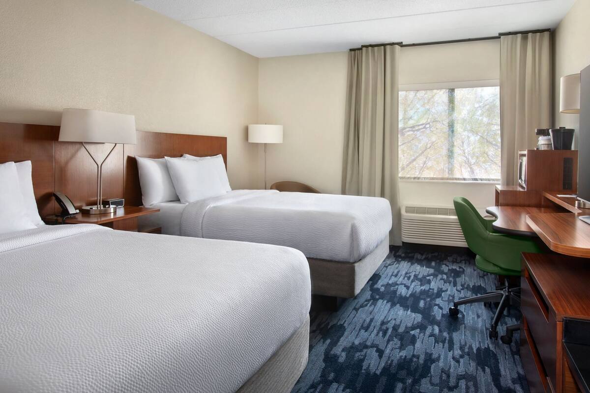 A hotel room with two beds, a desk, a chair, a lamp, and a window with curtains.