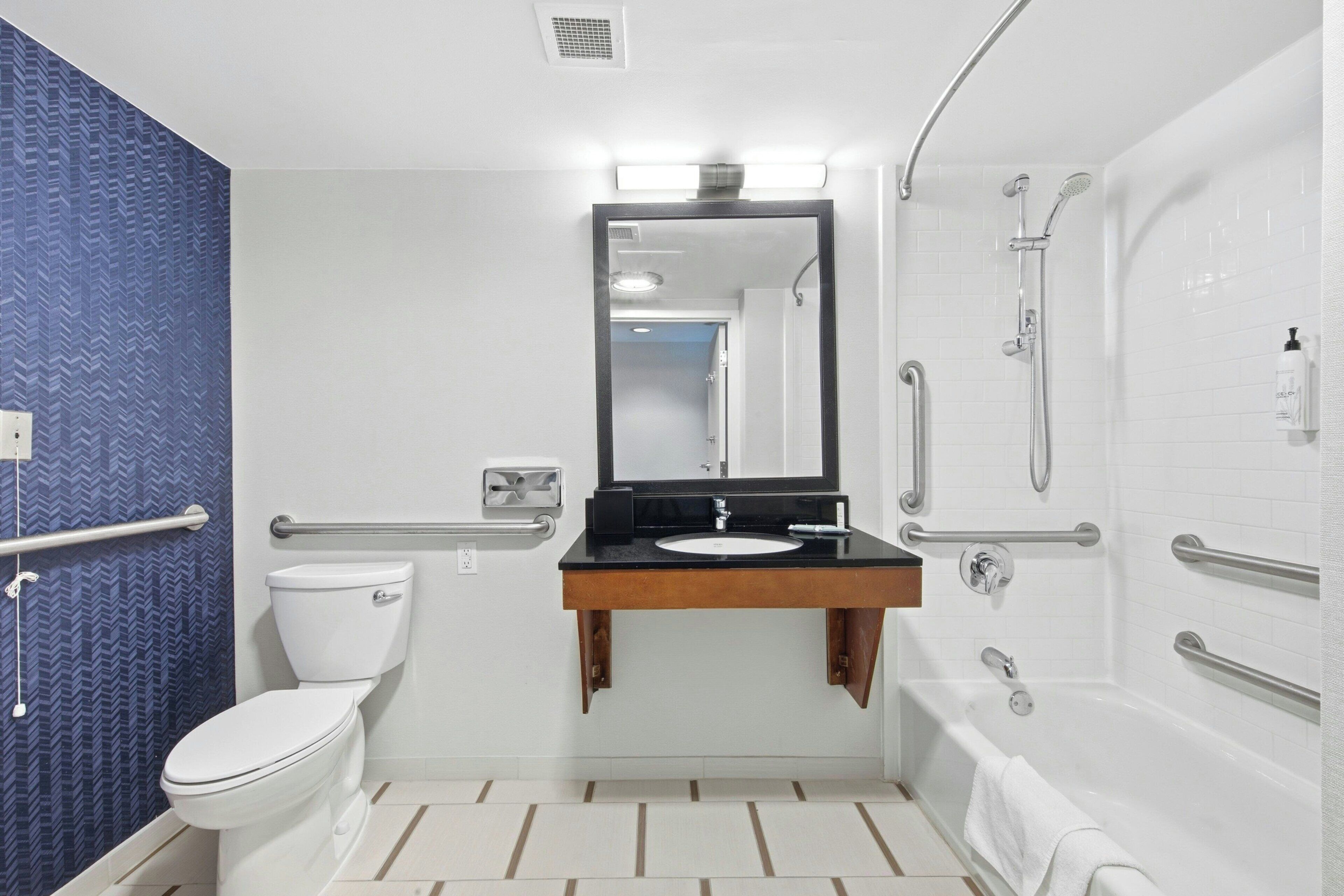 Een badkamer met een wit toilet, een wastafel met spiegel erboven, een douche met handgrepen en een betegelde vloer.