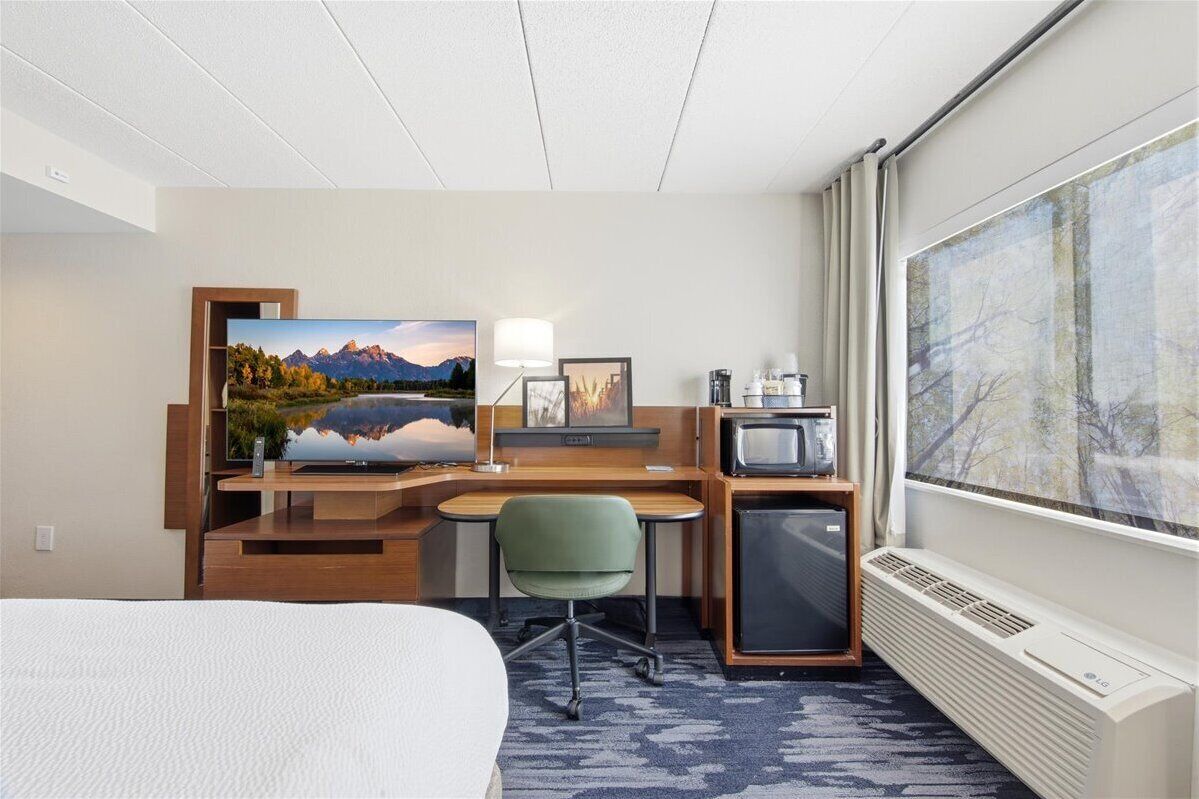 Habitación de hotel con cama, escritorio con computadora, televisor y ventana grande con vista a árboles.
