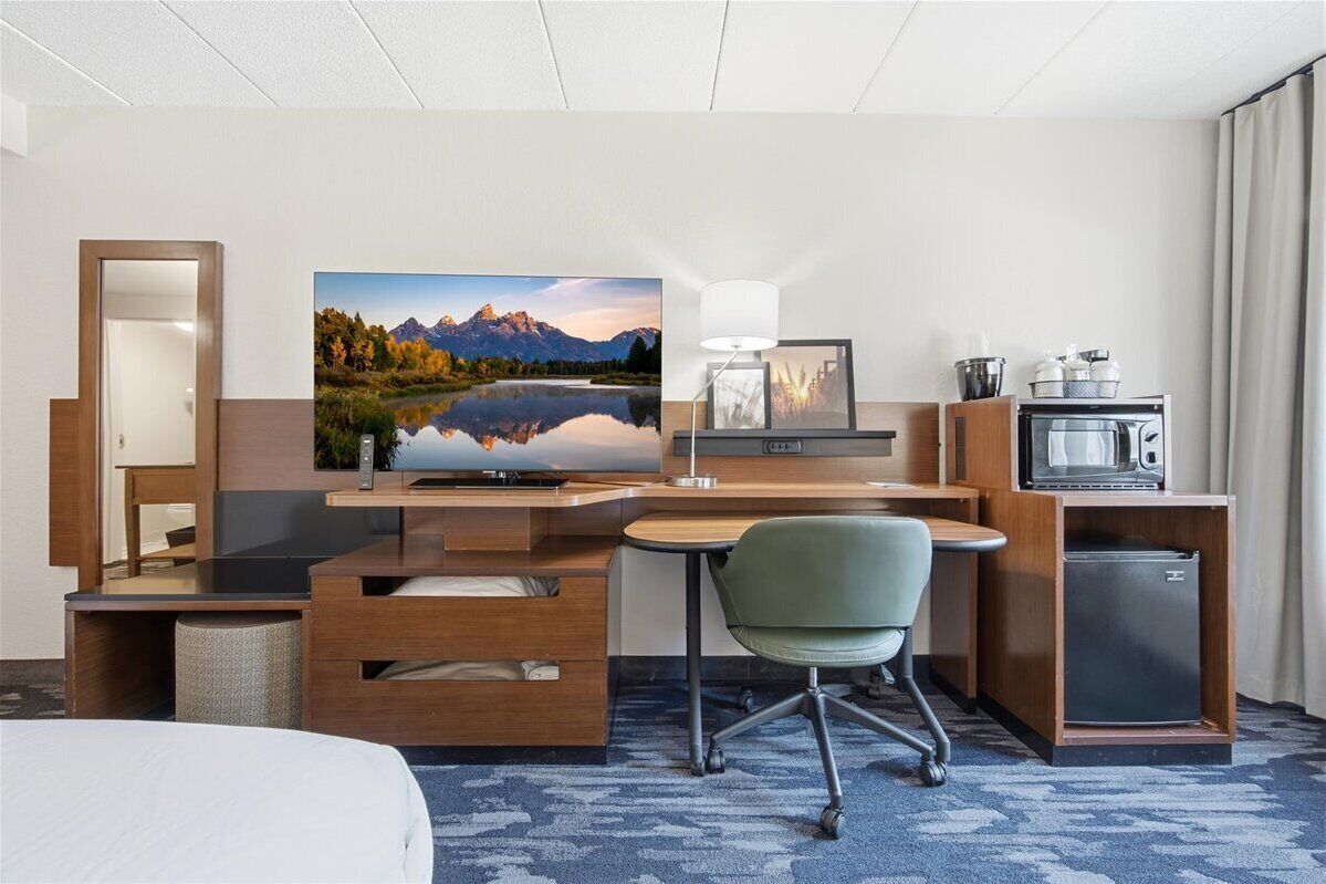 Ein Hotelzimmer mit einem Bett, einem Schreibtisch mit Computer, einem Fernseher, einer Lampe, einer Mikrowelle und einem kleinen Kühlschrank.