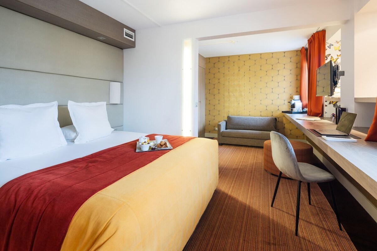 A hotel room with a bed, a desk with a laptop, a sofa, and a breakfast tray.