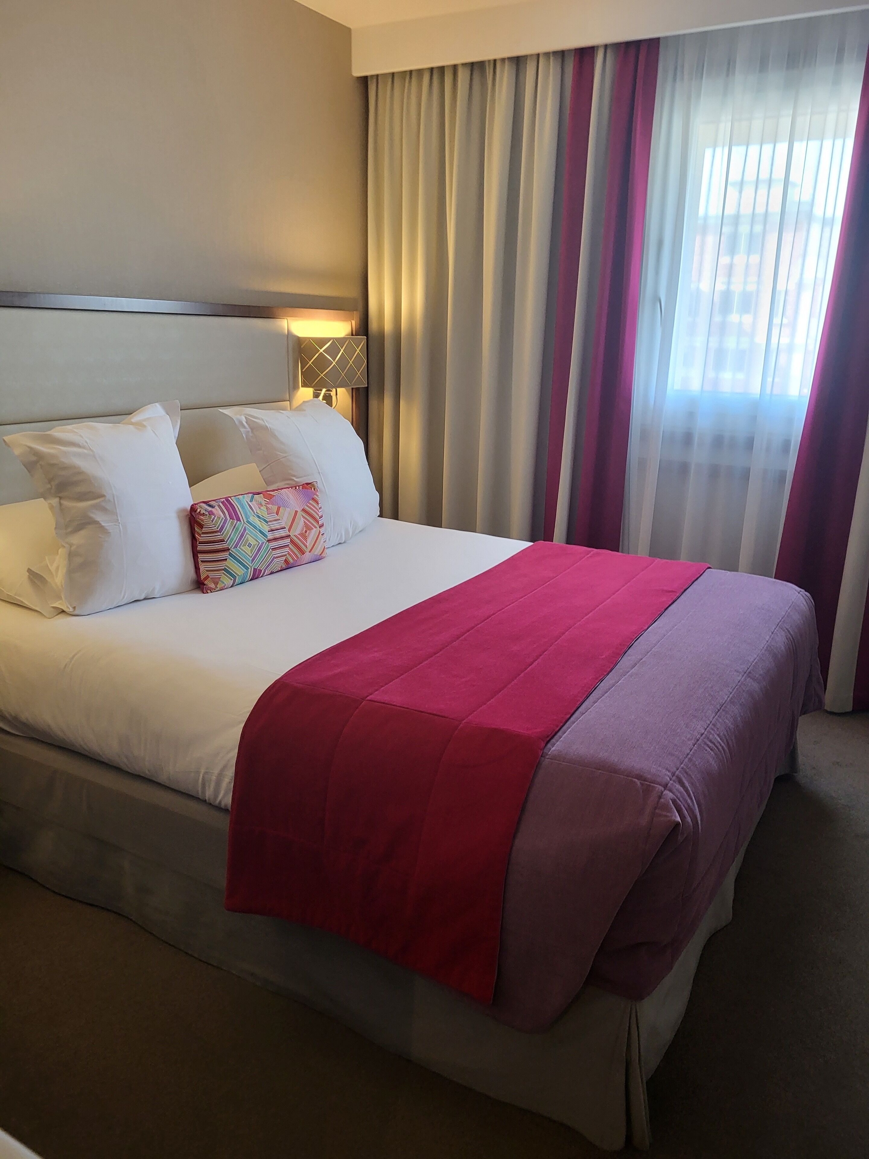 Een hotelkamer met een groot bed, wit beddengoed, een rode plaid en een raam met dunne gordijnen.