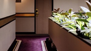 Un pasillo estrecho con alfombra morada, una puerta con número y una exhibición de plantas.