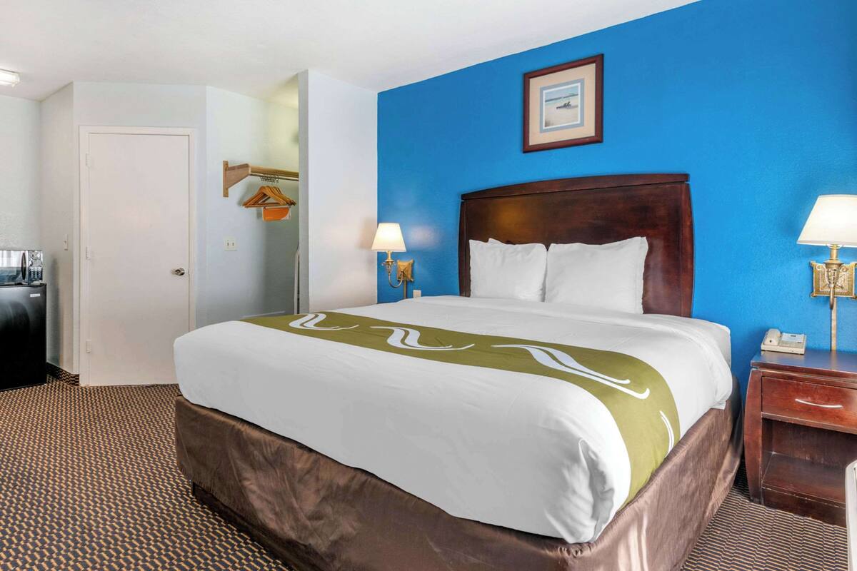 A hotel room with a large bed, two bedside lamps, a wooden nightstand, a framed picture on the wall, and a coat rack.