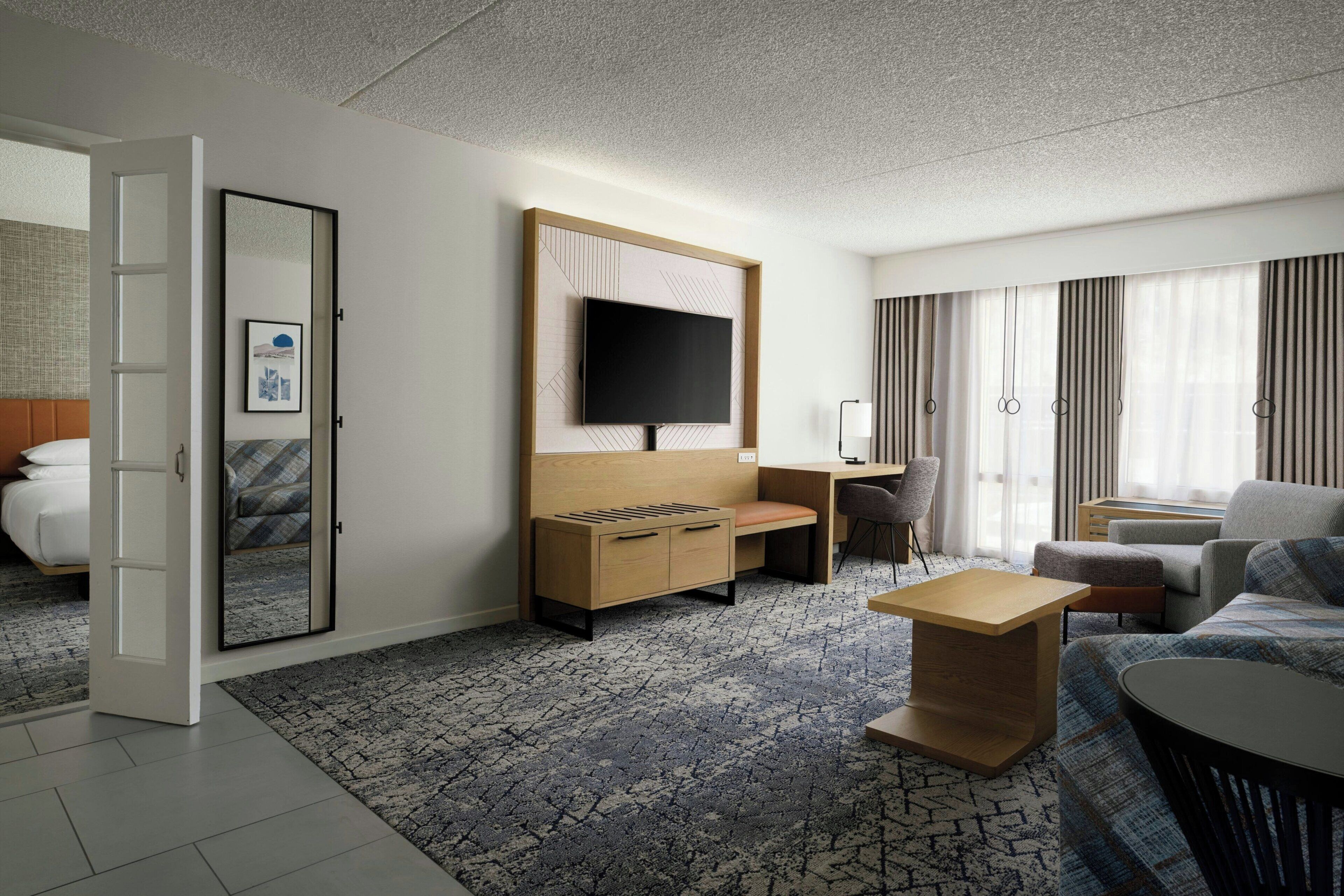 A modern hotel room with a bed, a desk, a television, and a seating area.