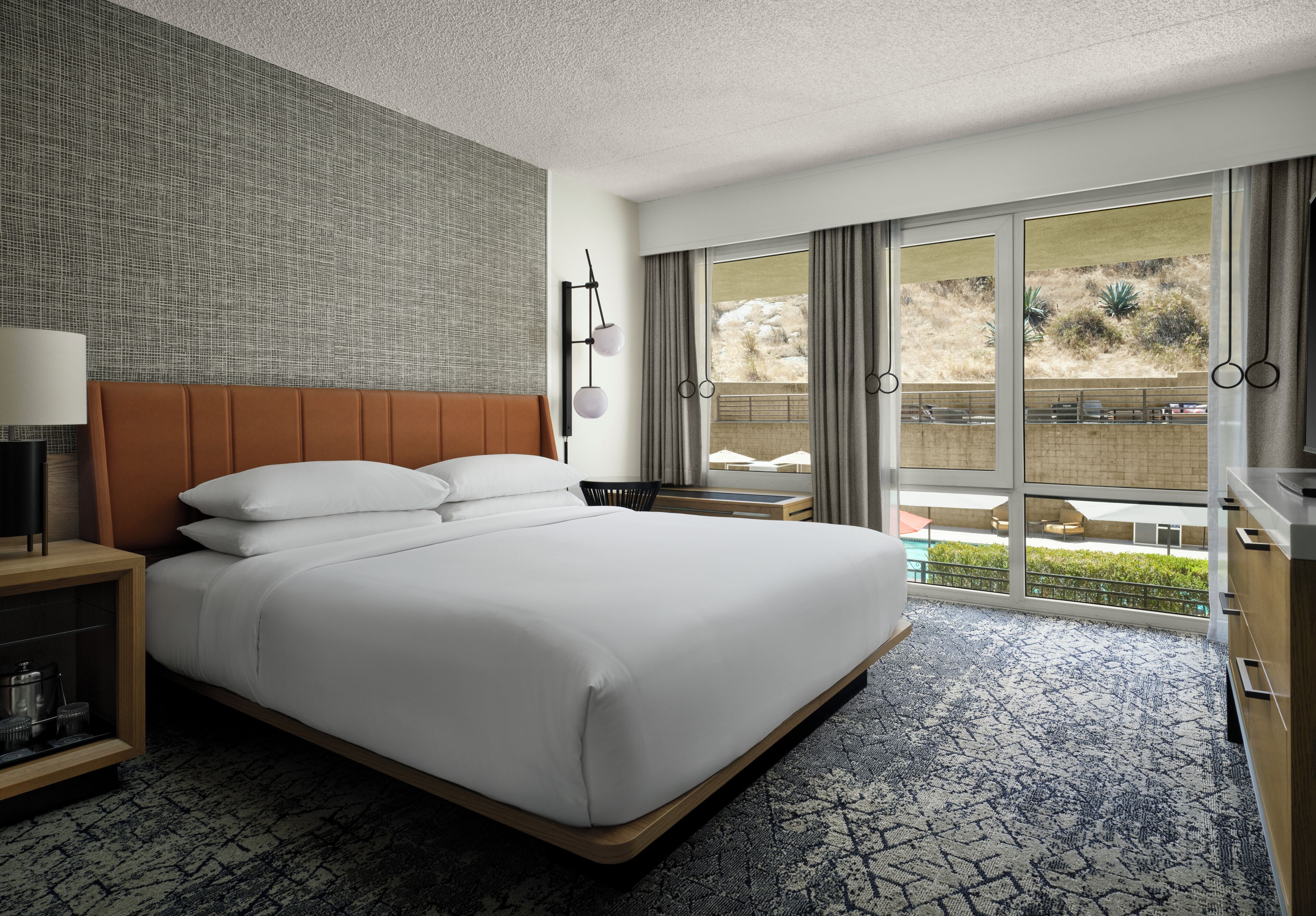 Habitación de hotel con una cama grande, una mesita de noche, una lámpara, un aplique de pared y vistas a una piscina y zona exterior.