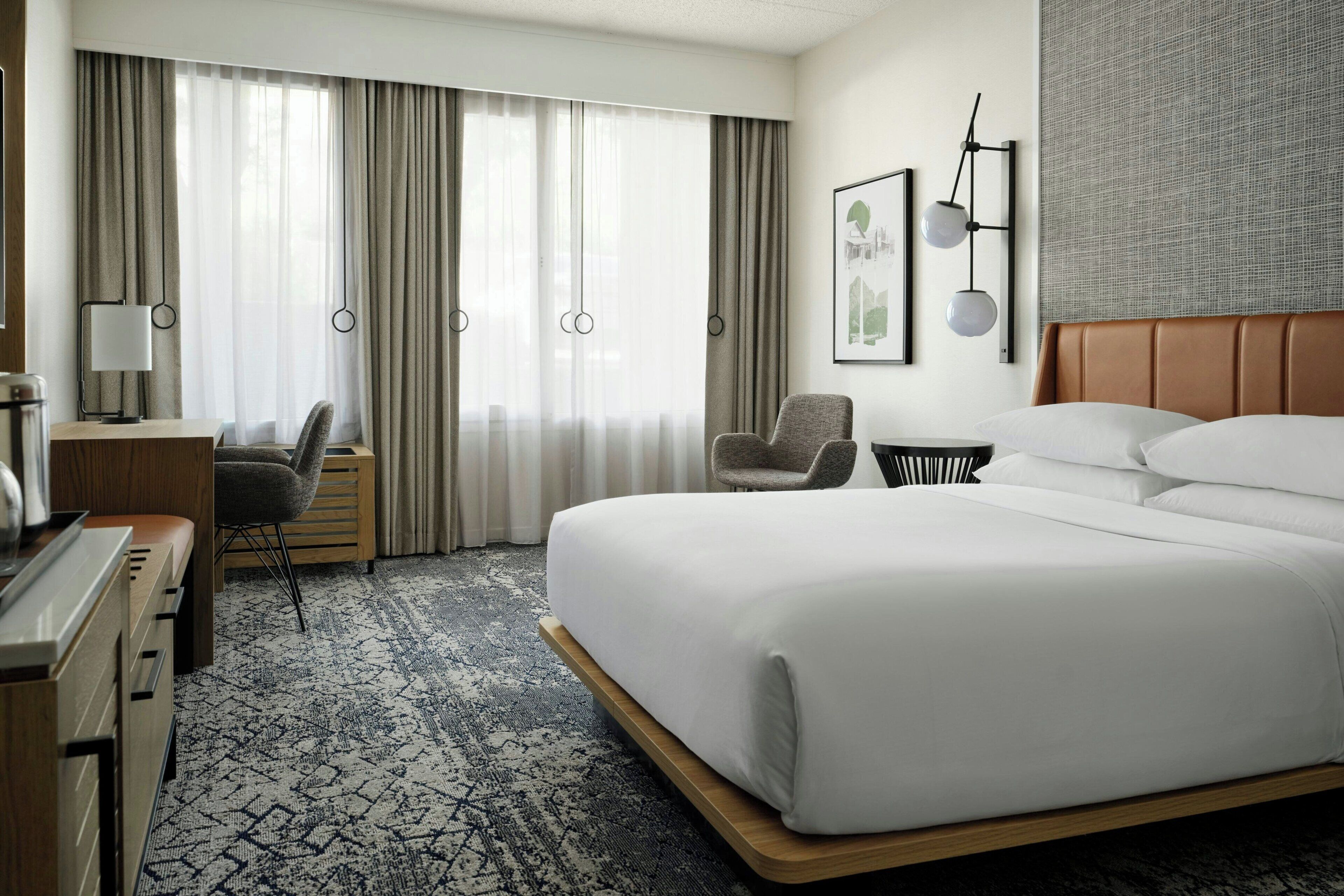 A hotel room with a large bed, a desk, a chair, a small table, and a wall-mounted lamp.