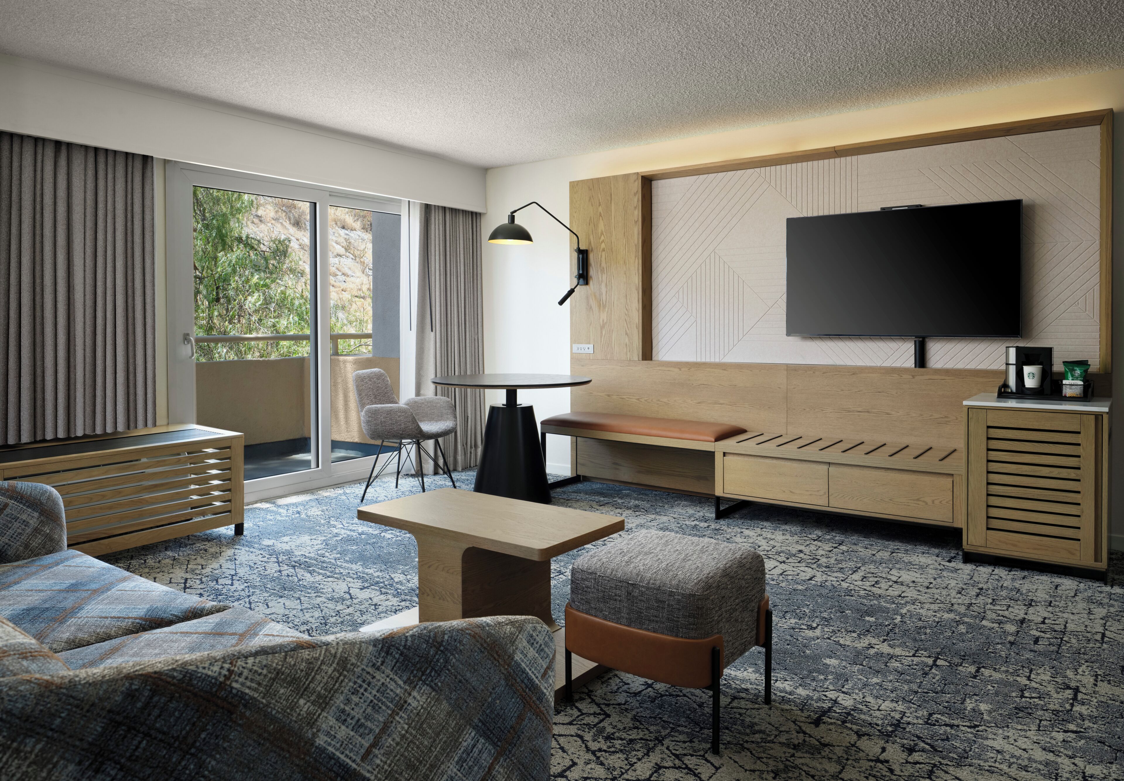 A modern hotel room with a sofa, armchair, a small table, a bench, a TV, and a coffee machine.