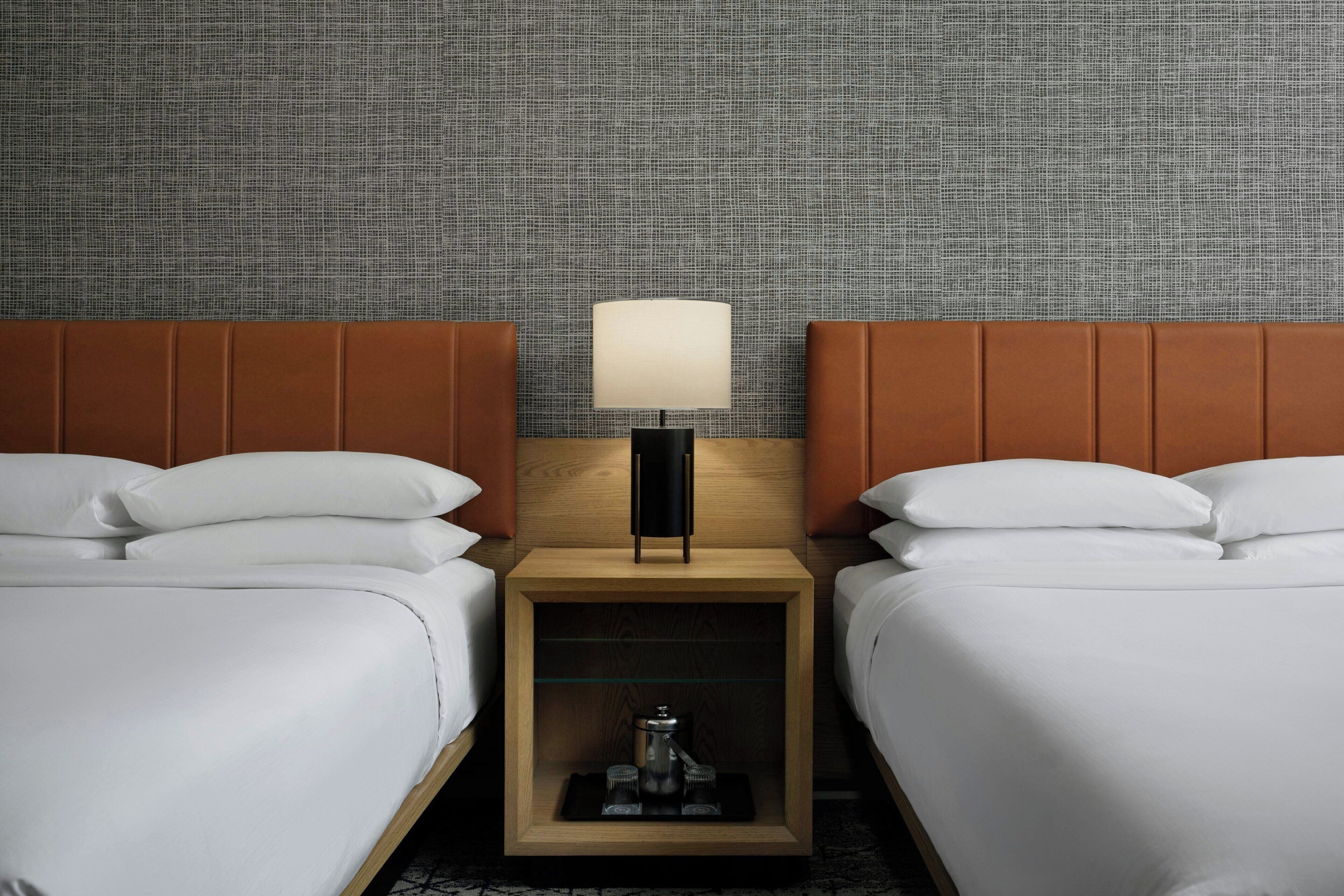 Habitación de hotel con dos camas, una mesita de noche con lámpara y un papel tapiz estampado.