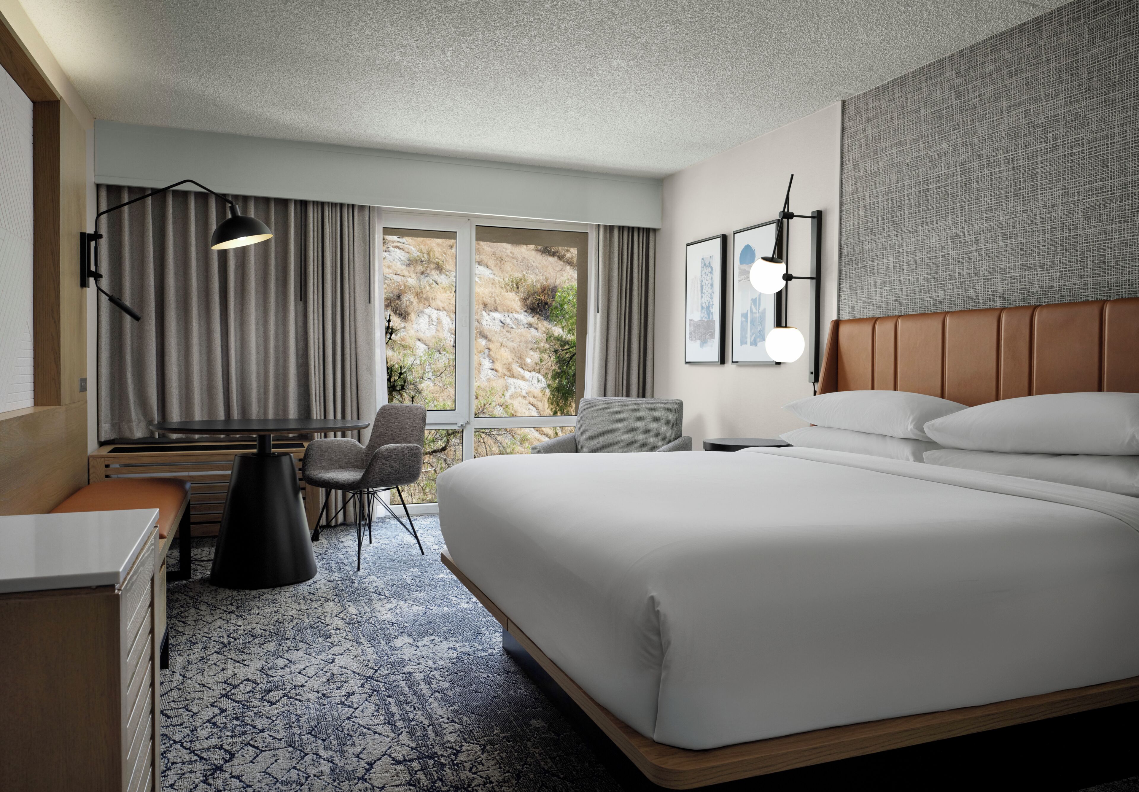 A modern hotel room with a large bed, a desk, a chair, and a view of a mountainous landscape through the window.
