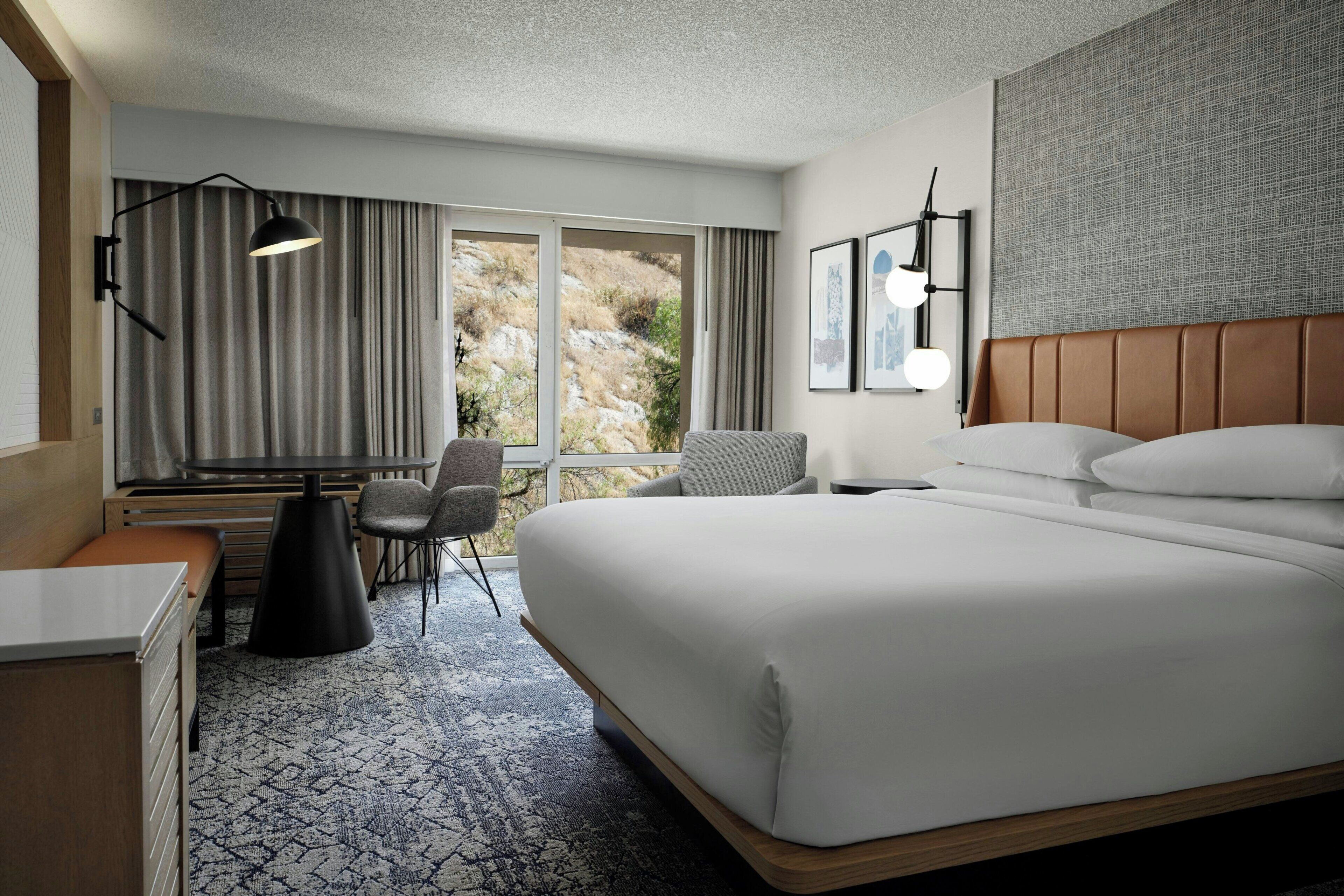 Habitación de hotel con una cama grande, un escritorio, una silla y vistas a un paisaje montañoso.