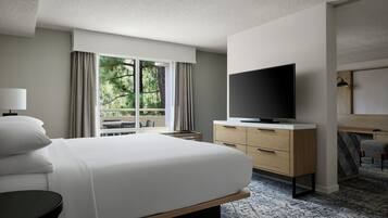 A hotel room with a large bed, a flat-screen TV, a desk, and a view of greenery through the window.