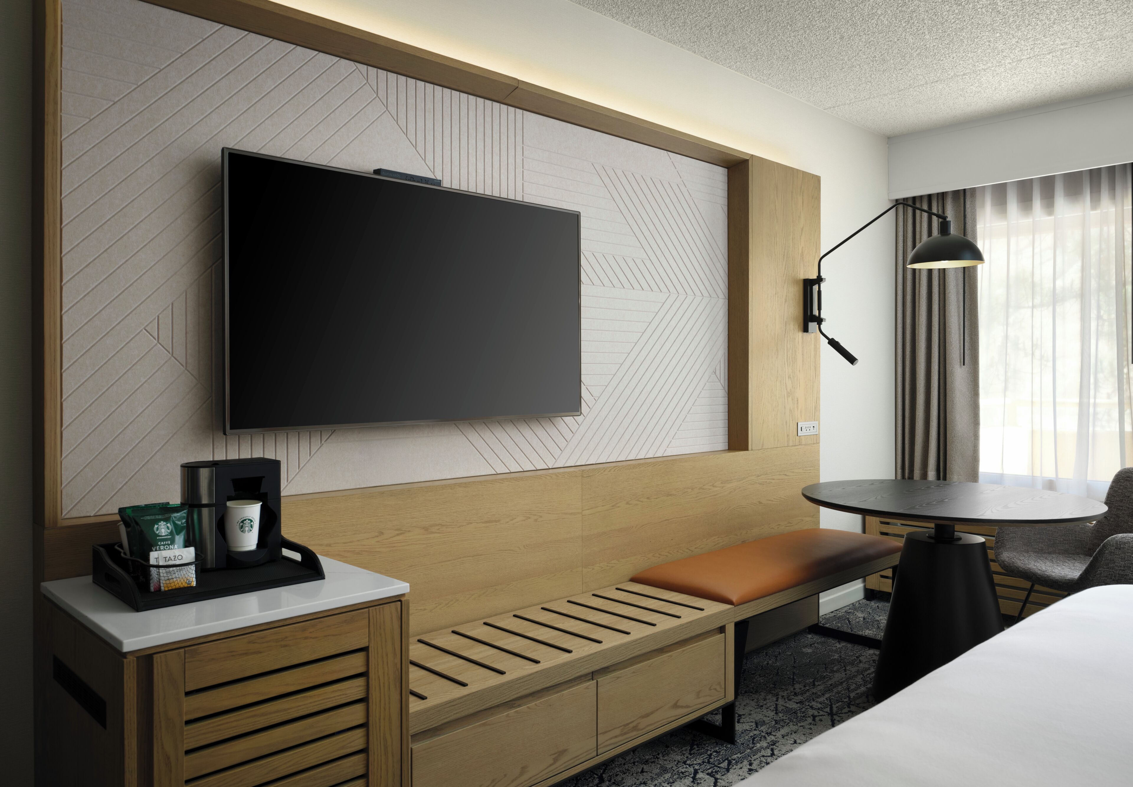Hotelkamer met een flatscreen televisie aan een structuurwand, een houten bankje, een klein tafeltje met een lamp en een koffieapparaat.