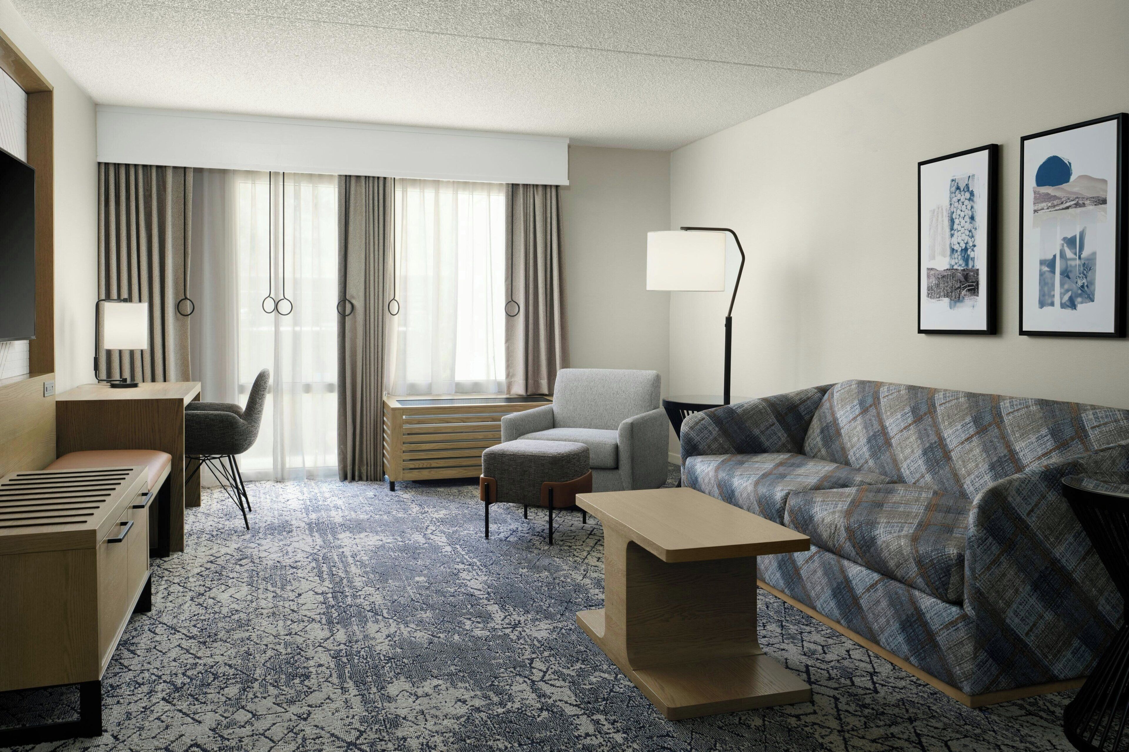 A hotel room with a sofa, armchair, desk, and a large window with curtains.