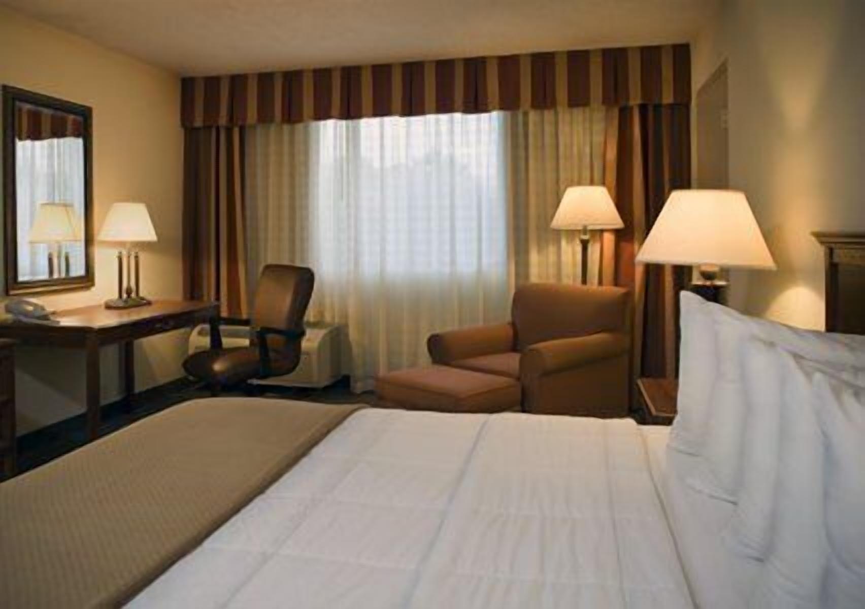 A hotel room with a large bed, two armchairs, a desk with a lamp, and a mirror.