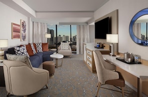 Habitación de hotel con sofá, sillones, zona de comedor, televisor y vistas a la ciudad.