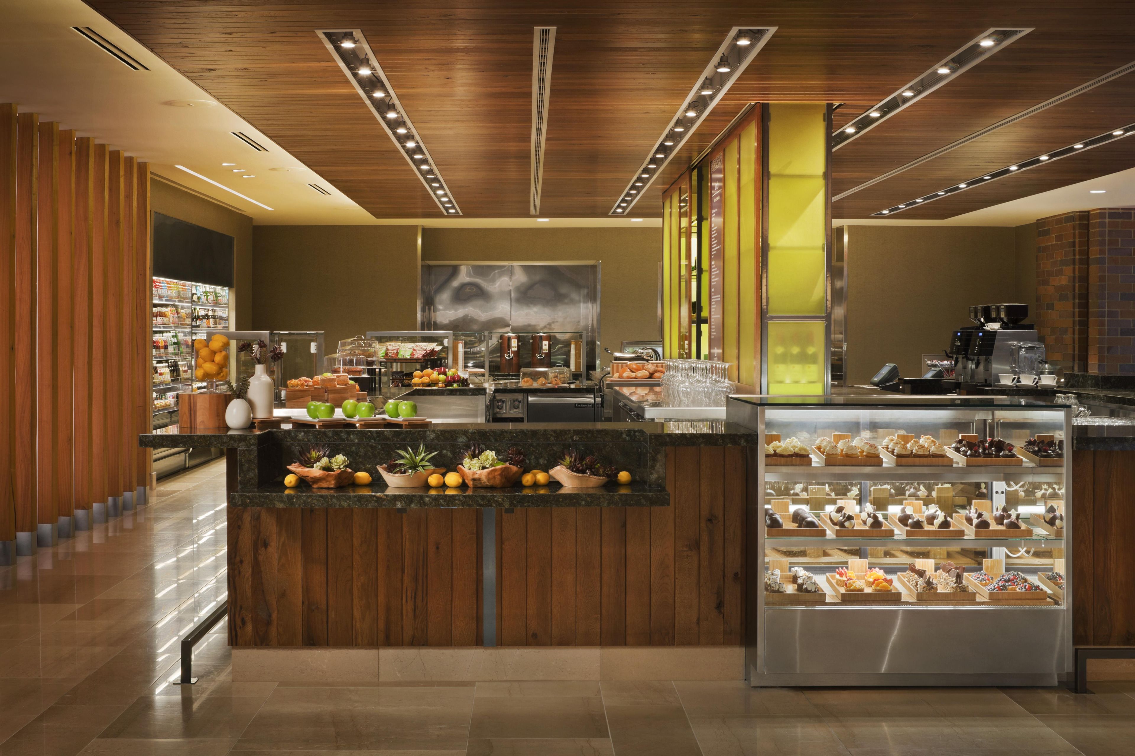 A modern cafe with a wooden interior, a central counter with fresh fruits and pastries, and a display case with desserts.