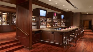 A bar area with wooden flooring, a long counter with stools, and shelves stocked with bottles and glasses.