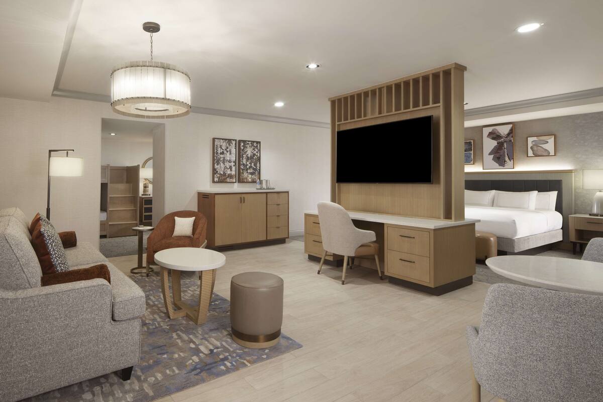 A modern hotel room with a sofa, a desk, a chair, a TV, a bed, and a bedside table.