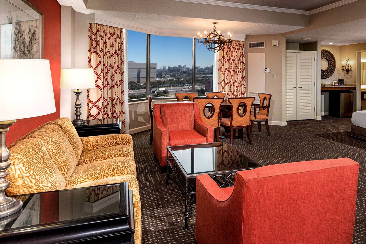 A hotel room with a sofa, armchair, dining table, chairs, and a view of the cityscape through large windows.