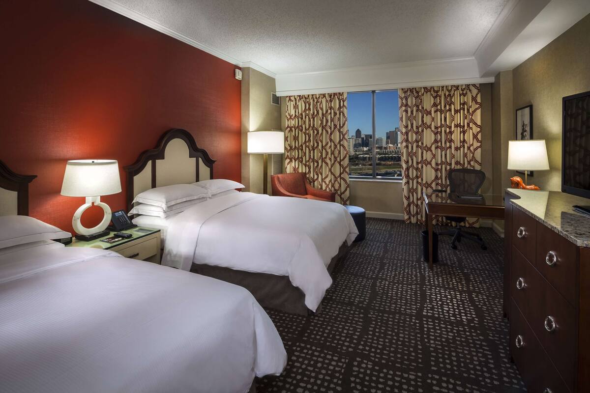 A hotel room with two beds, a desk, a chair, and a view of the city skyline.