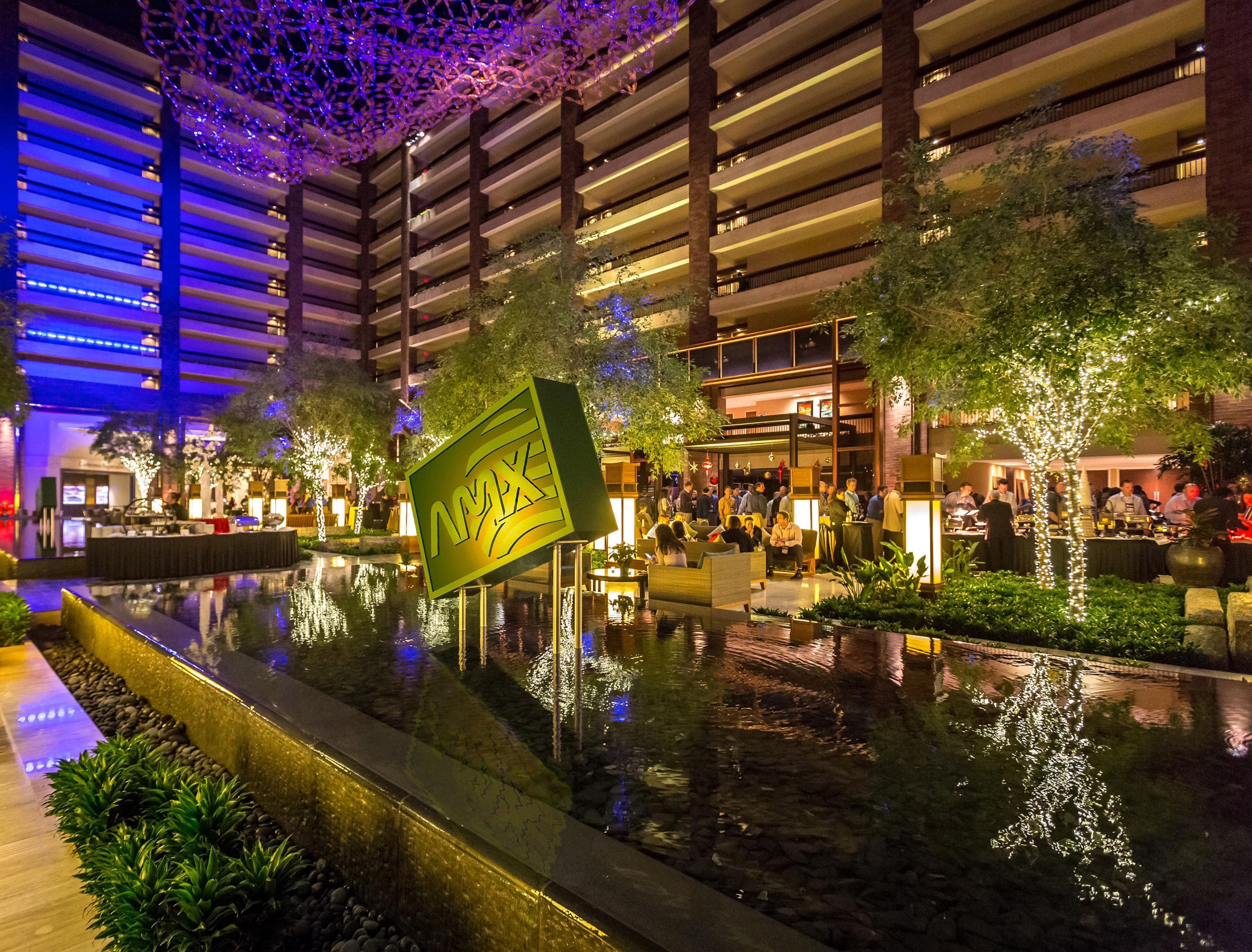 A modern indoor atrium with a reflective water feature, seating areas, and a large illuminated sign.