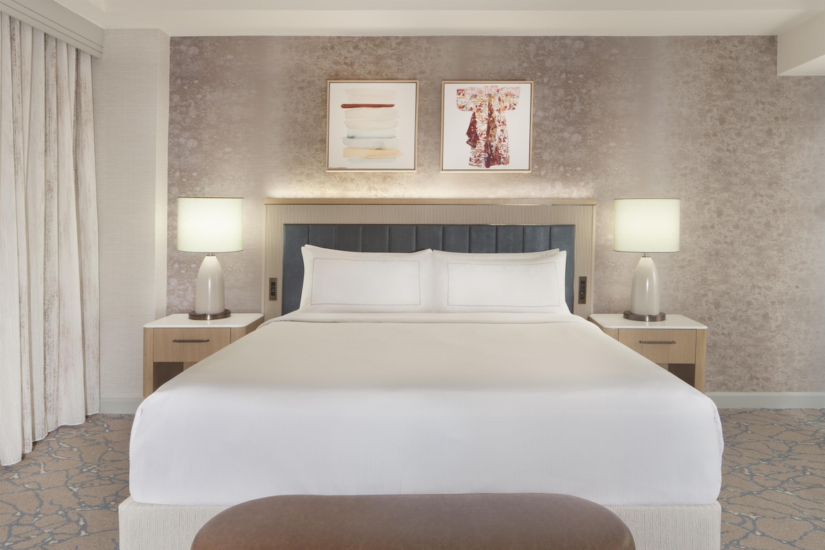 A hotel room with a large bed, two bedside tables with lamps, a headboard, and two framed artworks on the wall.
