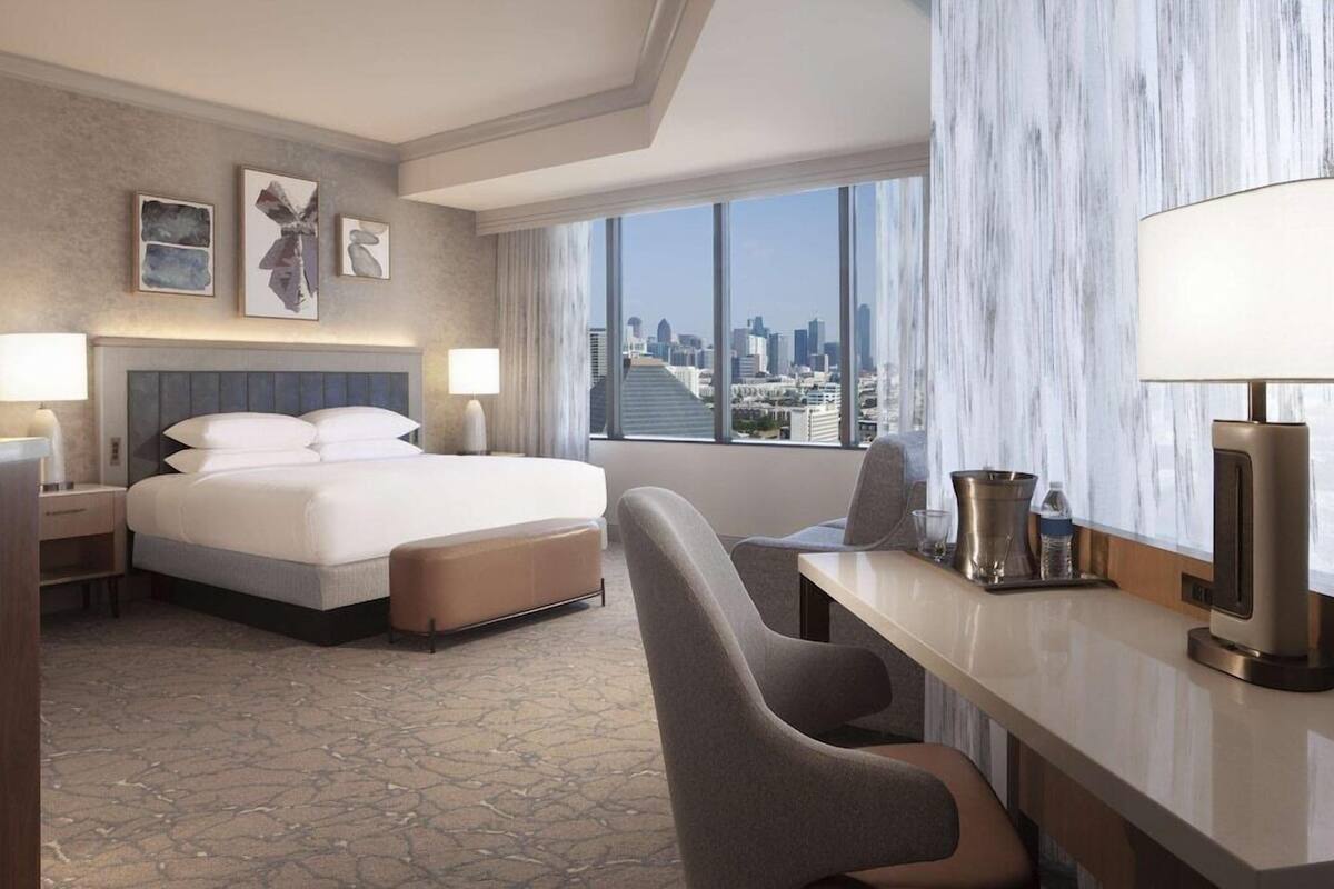 A hotel room with a large bed, a desk with a lamp, a chair, and a view of the city.