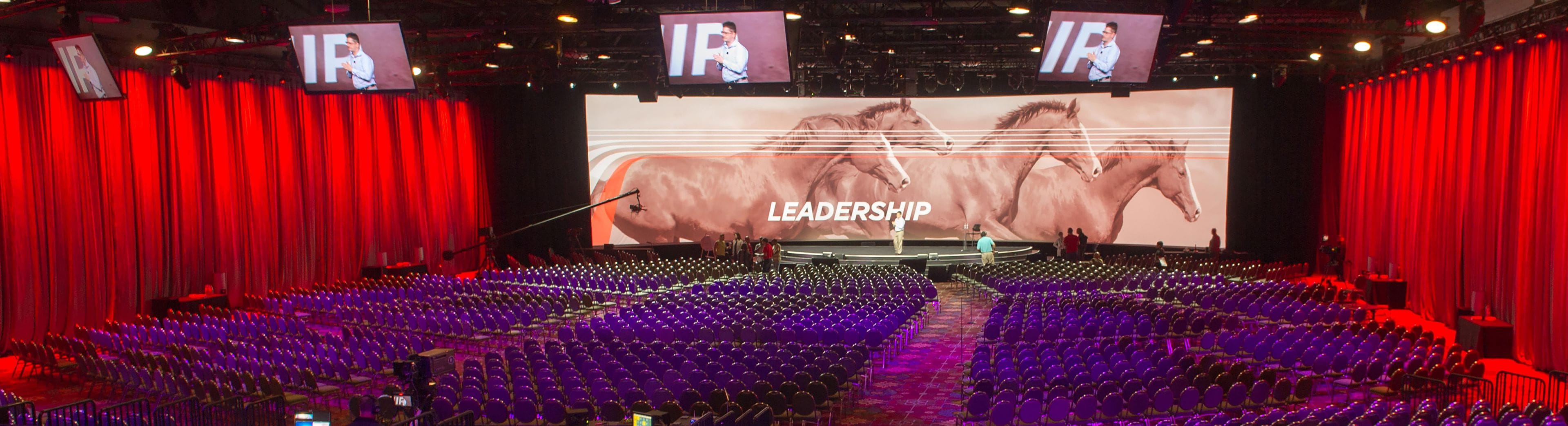 A large auditorium with rows of purple chairs, a stage with a large screen displaying horses and the word 'LEADERSHIP,' and red curtains.