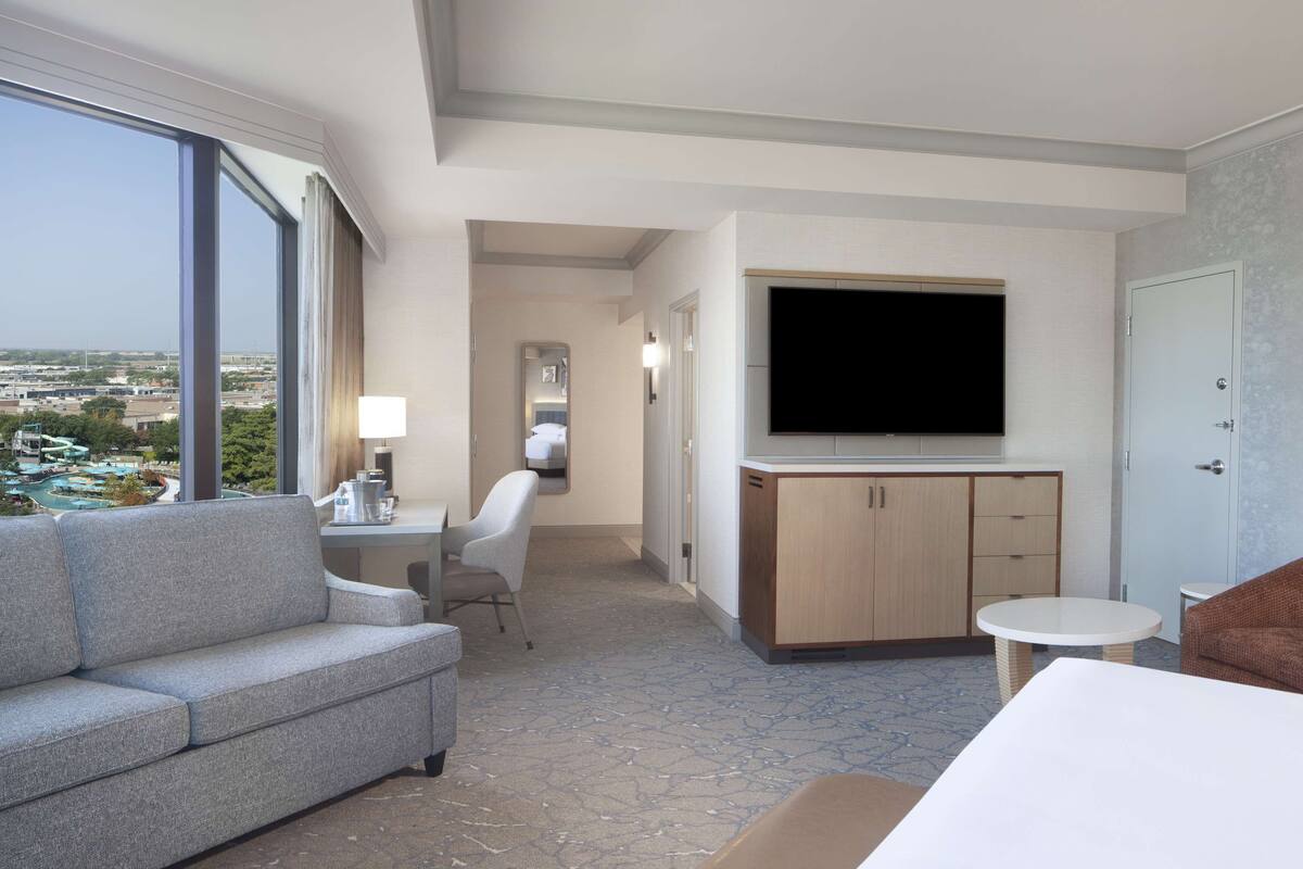 A modern hotel room with a large window, a flat-screen TV, a sofa, a small table, and a chair.
