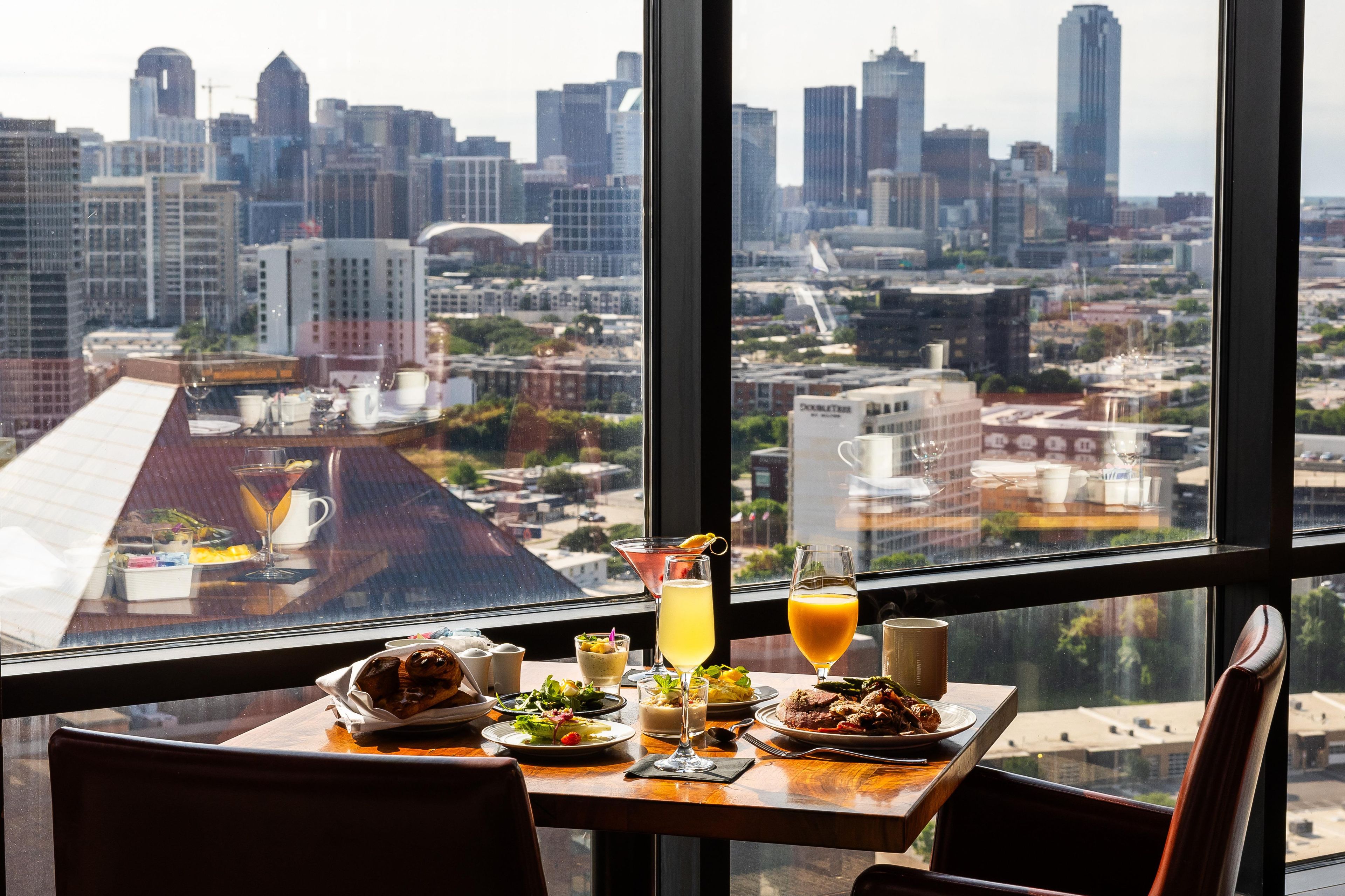 A dining table with a meal and drinks, a city view through large windows, and a chair.