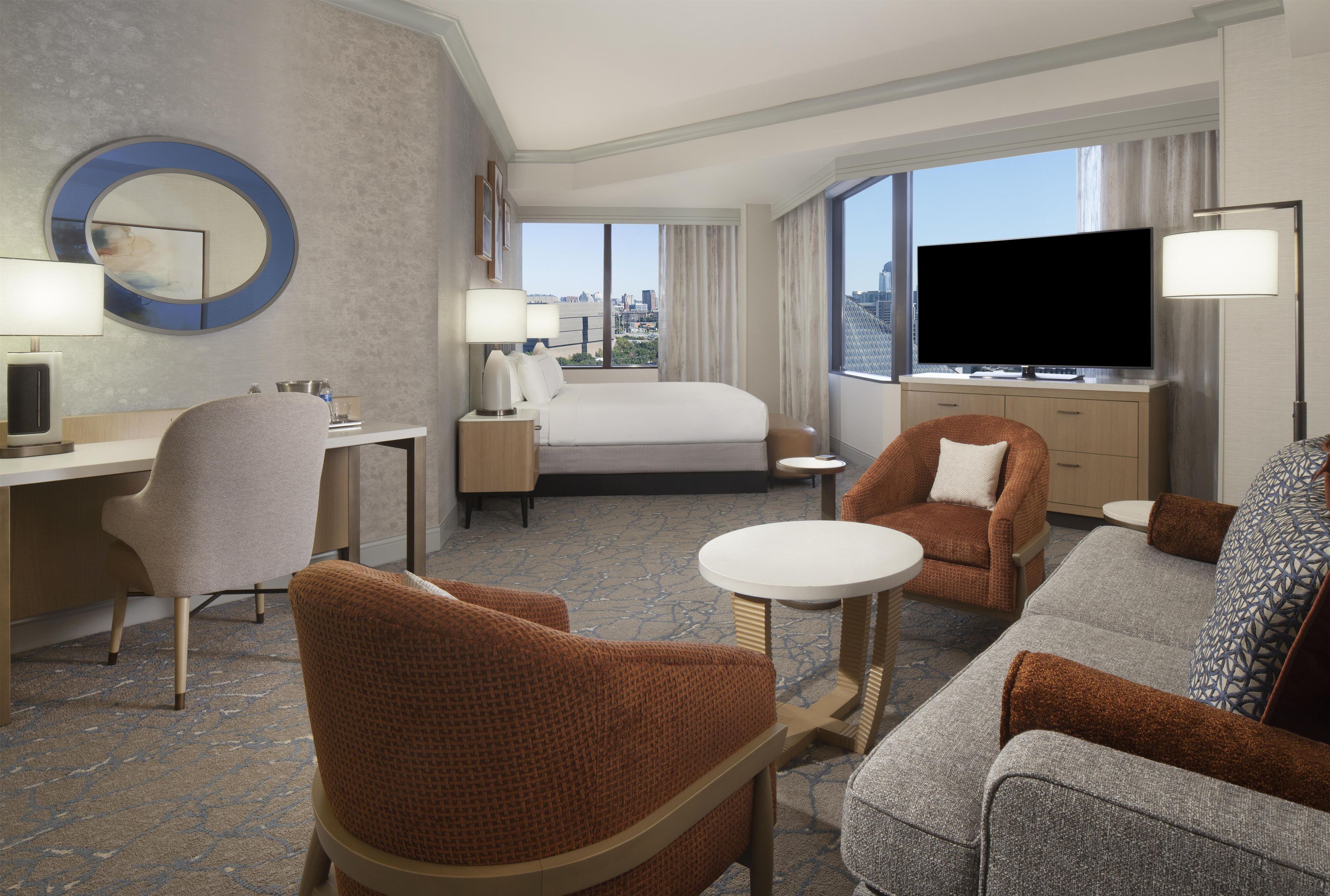 A hotel room with a bed, two armchairs, a sofa, a desk, a television, and a large window with a city view.