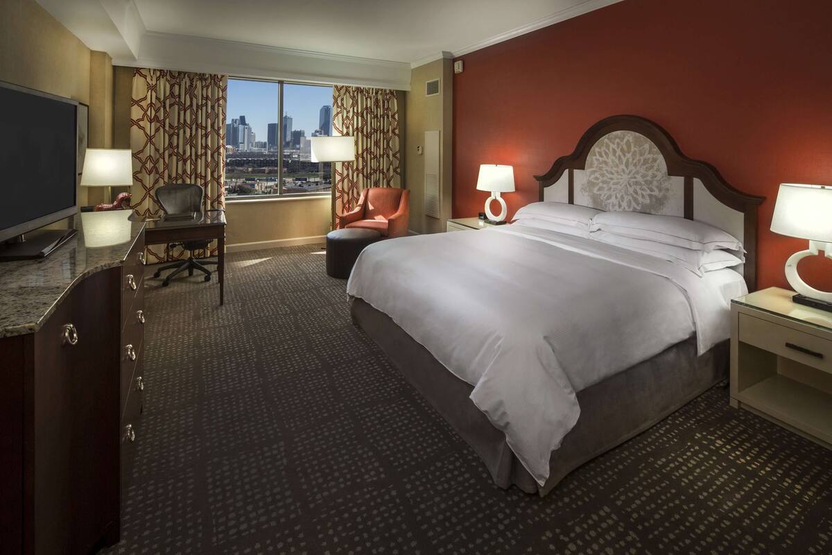 A hotel room with a large bed, a desk with a TV, a chair, and a view of a cityscape.