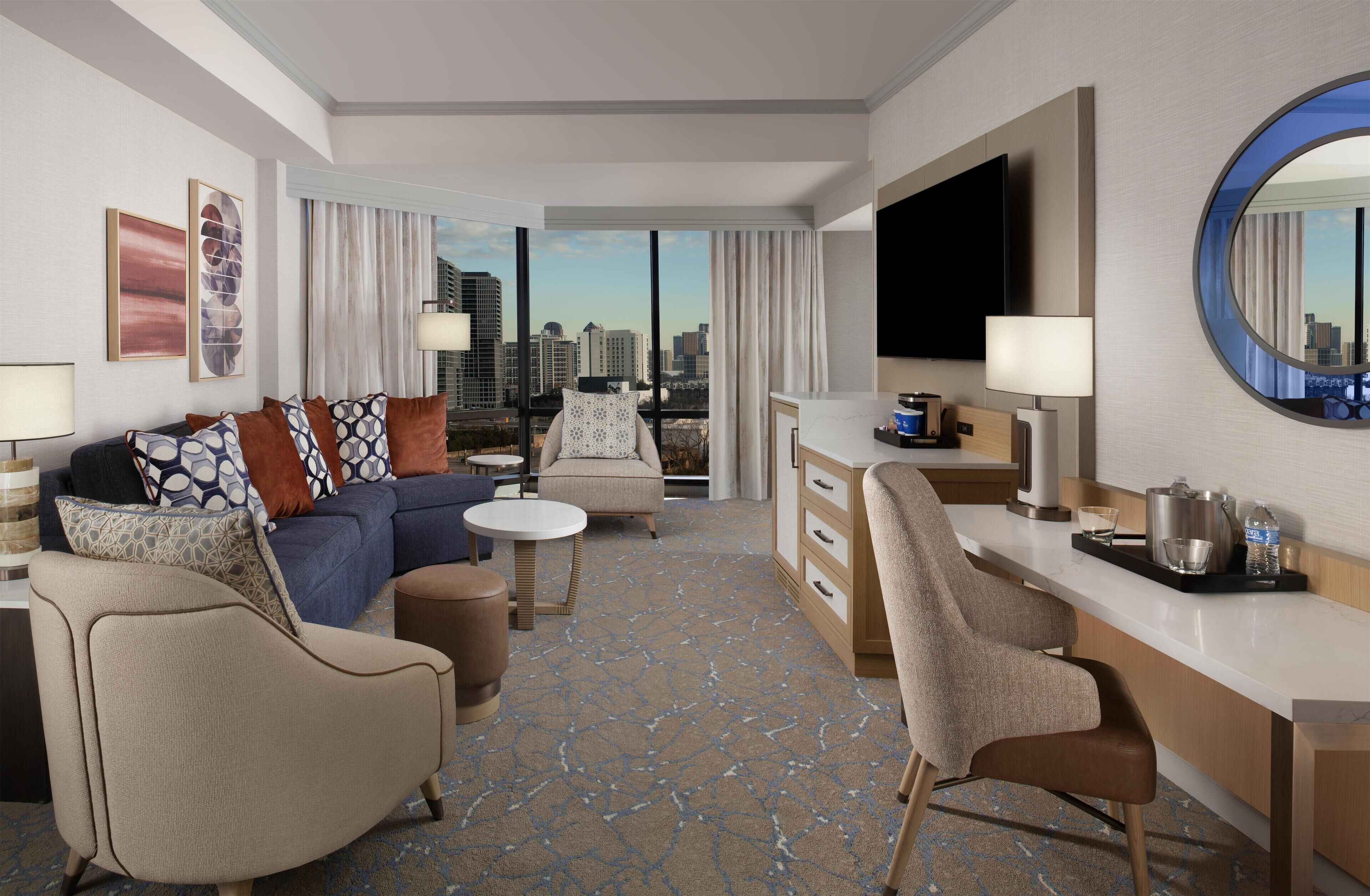 A modern hotel room with a sofa, armchairs, a dining table, a TV, and a city view.