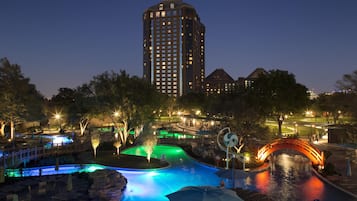 A nighttime view of a hotel with a swimming pool, a bridge, and a tall building in the background.
