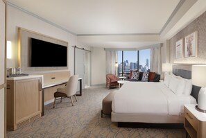 A hotel room with a large bed, a desk with a flat-screen TV, a chair, a small table, and a view of the city skyline.