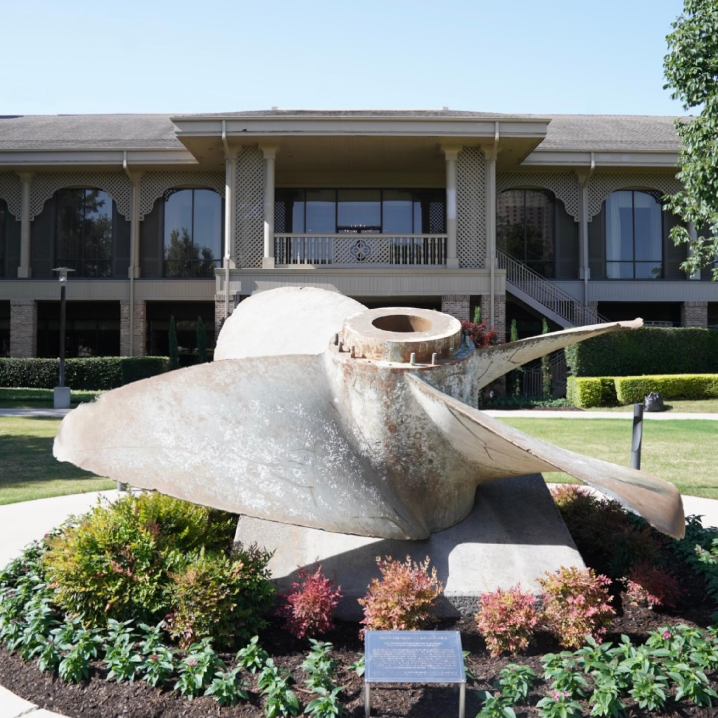 Une grande sculpture représentant un poisson, située dans un espace paysager, avec un bâtiment en arrière-plan.