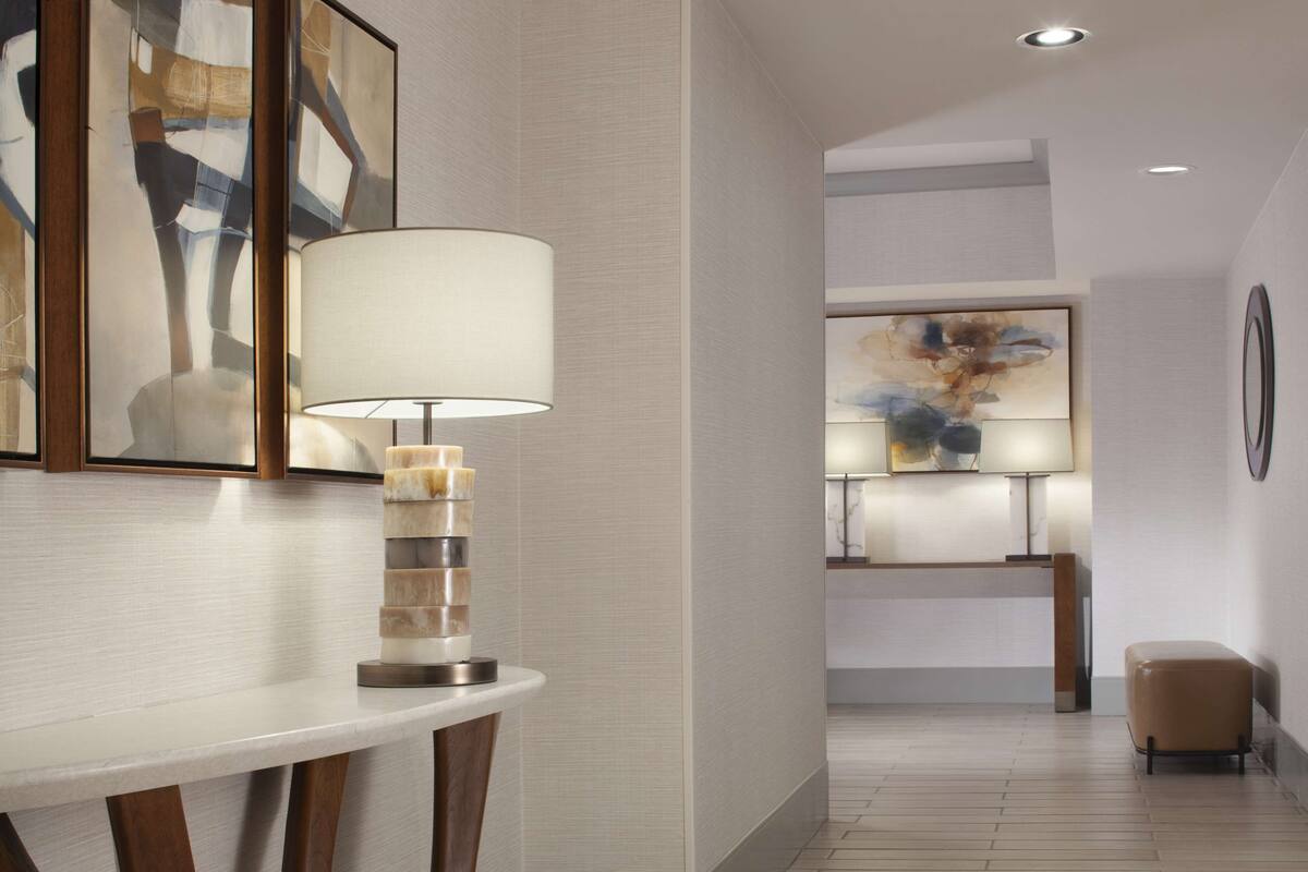 A modern hotel lobby with a minimalist design, featuring a round table with a unique lamp, framed artwork on the walls, and a wooden floor.