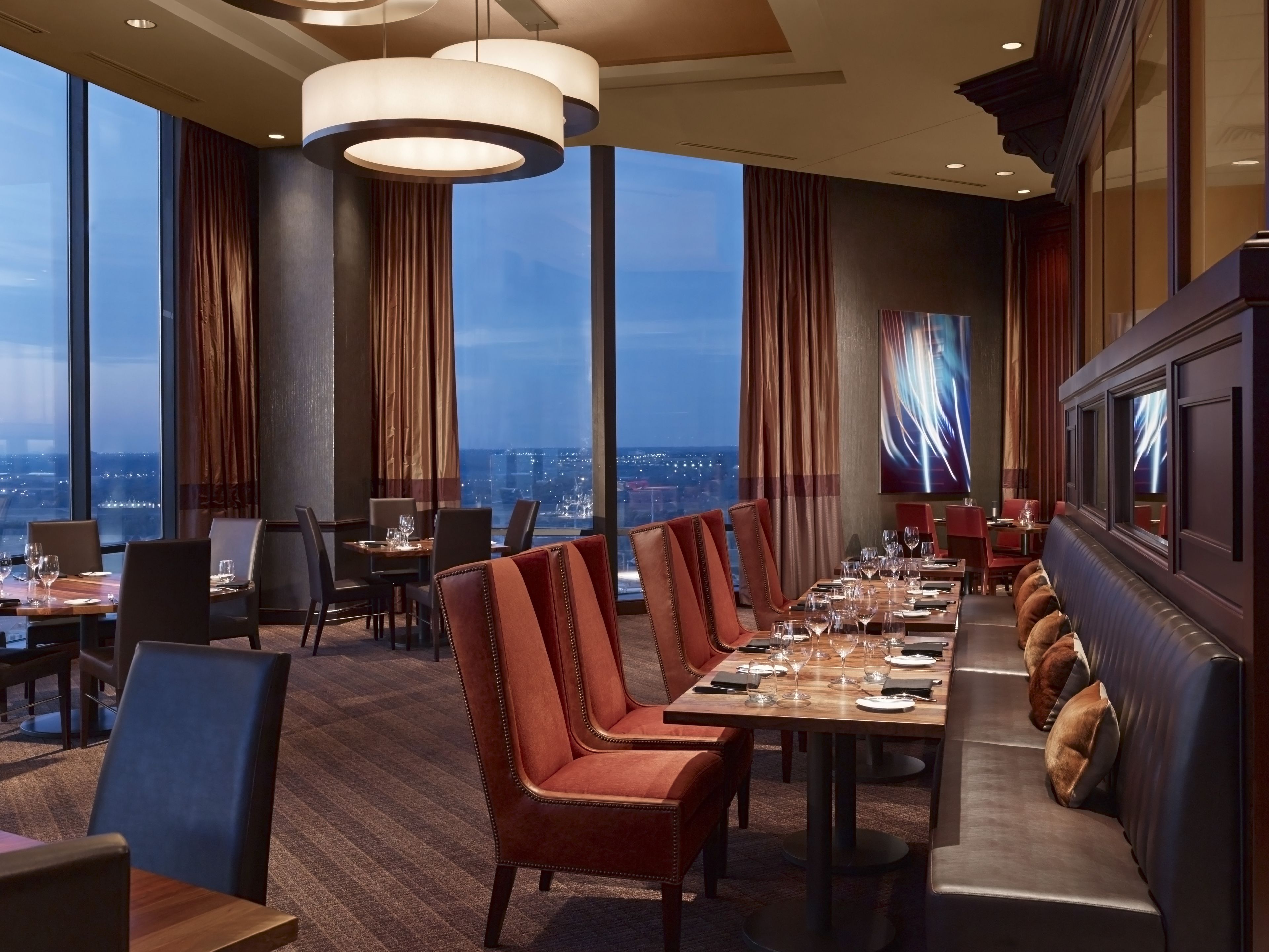 A modern dining area with high tables and leather chairs, large windows with curtains, and a city view.