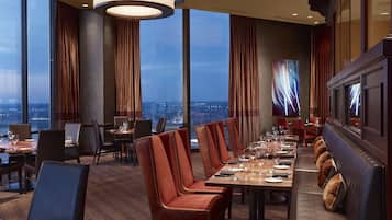 A modern dining area with high tables and leather chairs, large windows with curtains, and a city view.