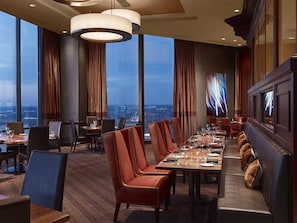 A modern dining area with high tables and leather chairs, large windows with curtains, and a city view.