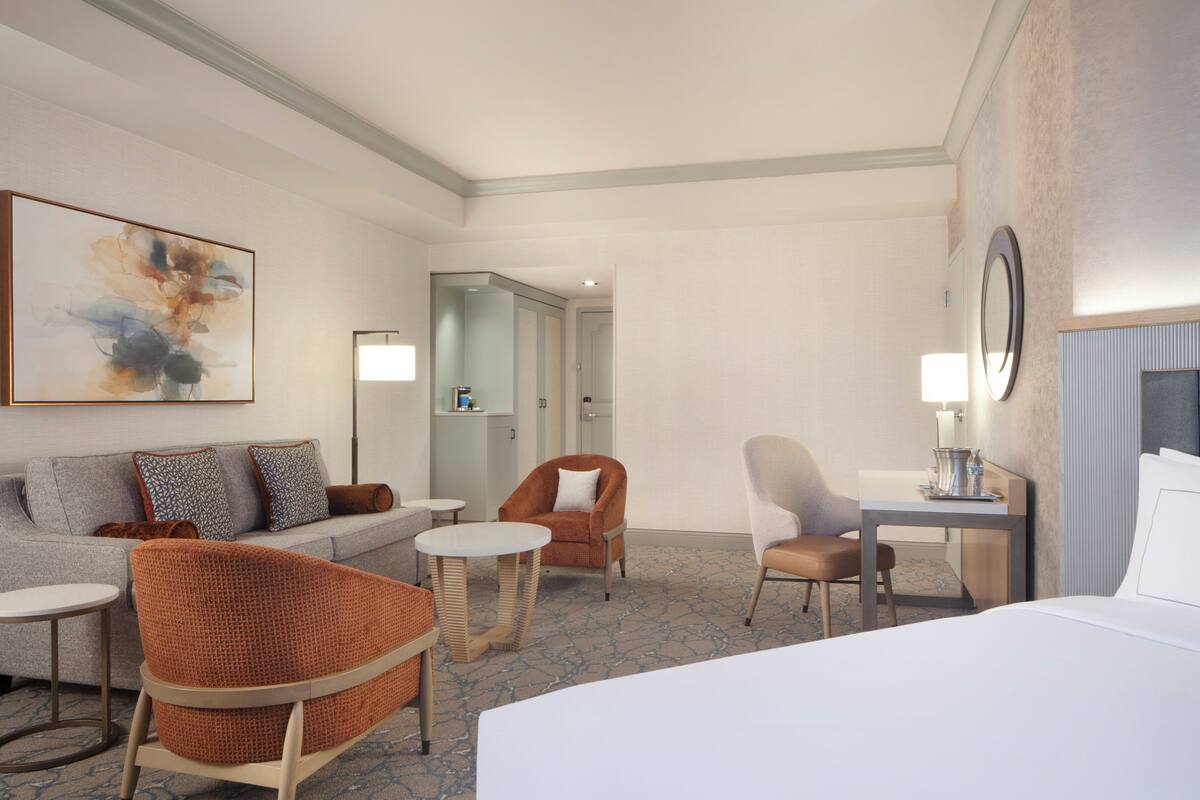 A hotel room with a bed, a sofa, two armchairs, a small table, a desk with a lamp, a mirror, and a painting on the wall.