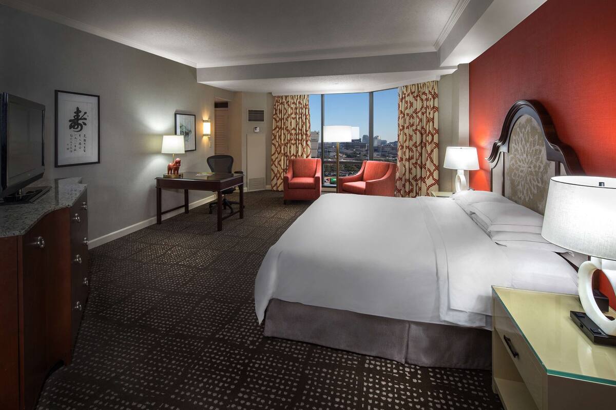 A hotel room with a large bed, two armchairs, a desk, a TV, and a view of the city.