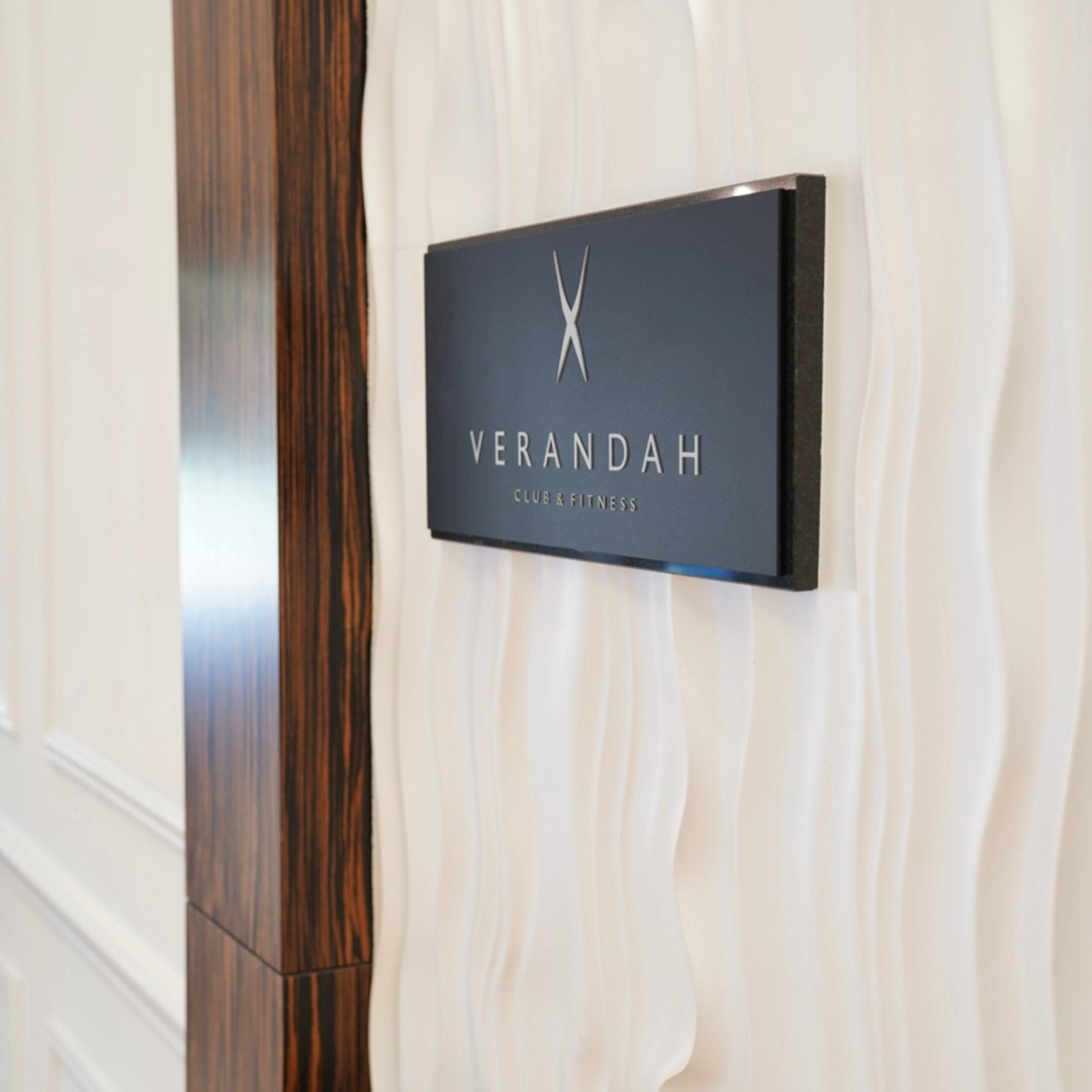 A black sign with the word 'VERANDAH' and the words 'CLUB & FITNESS' on a textured wall.