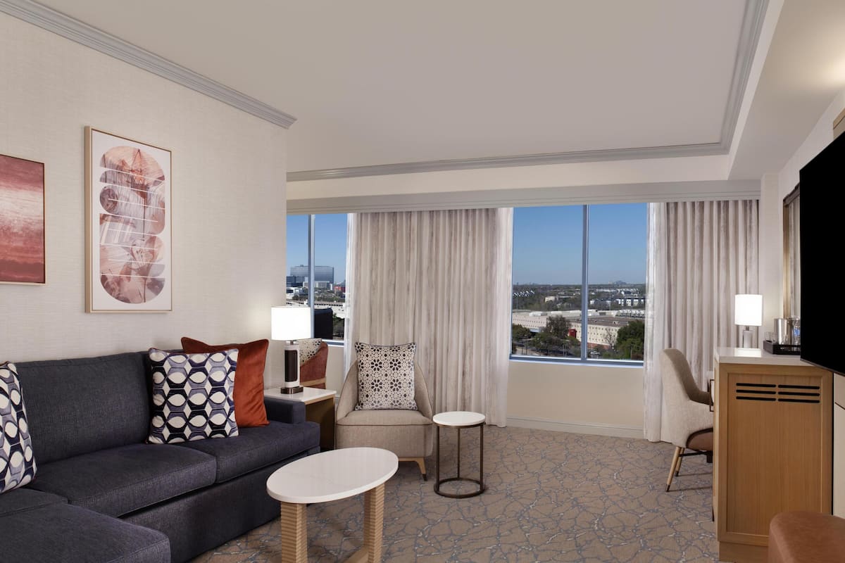 A modern hotel room with a sofa, two chairs, a small table, a TV, and a large window with curtains.