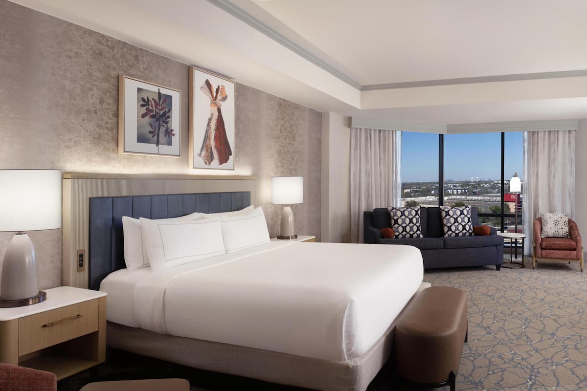 A hotel room with a large bed, a sofa, a chair, a nightstand, a lamp, and a view of the city.