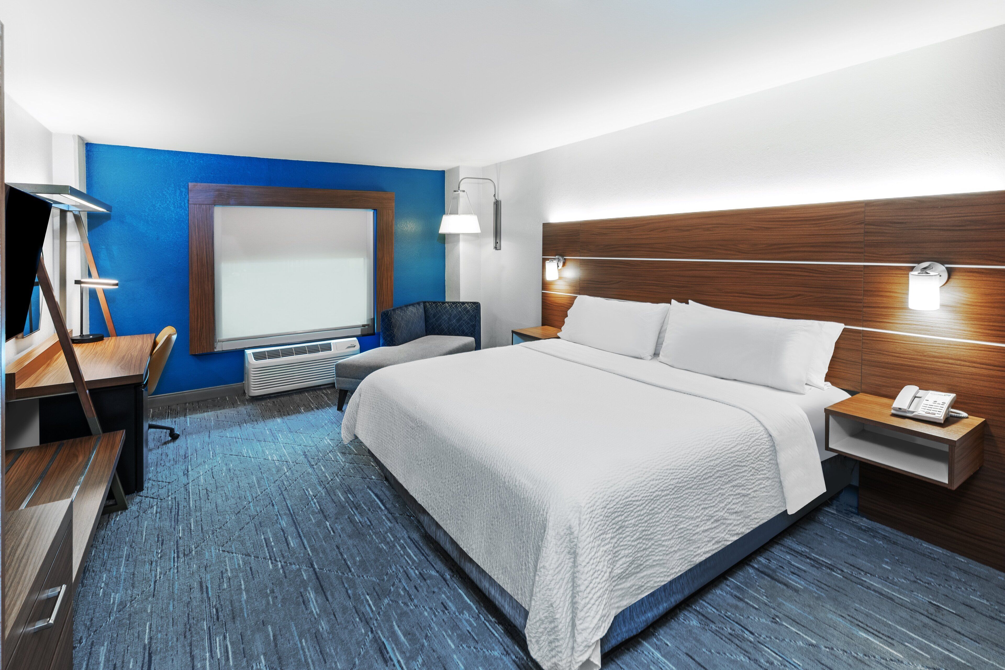 A hotel room with a large bed, a desk, a chair, a desk lamp, a nightstand with a phone, and a wall-mounted lamp.