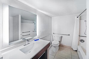 Un baño moderno con lavamanos, espejo y retrete blancos. Cuenta con una zona de ducha con puerta de cristal y una barra de apoyo para seguridad.